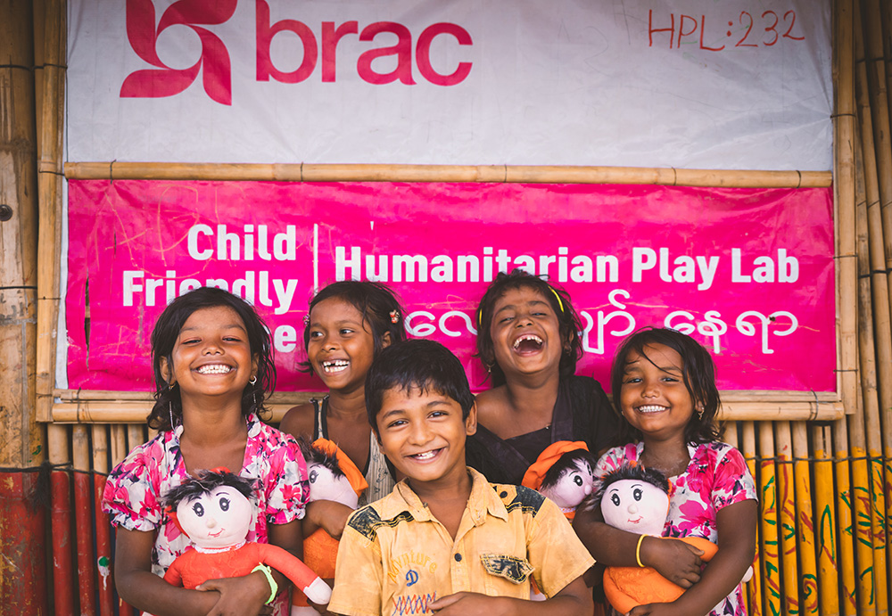 A group of five children holding dolls smile and laugh as they stand in front of a sign for a BRAC Humanitarian Play Lab