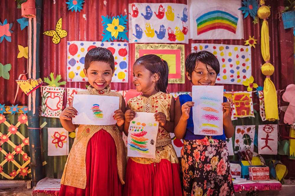 Three children in a Play Lab in Bangladesh hold up their colorful paintings and smile