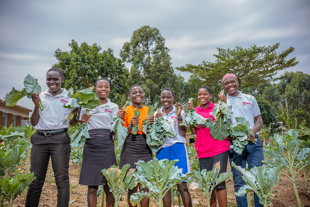A group of six young men and women hold up produce from their school garden that they started through a BRAC ELA club in Uganda