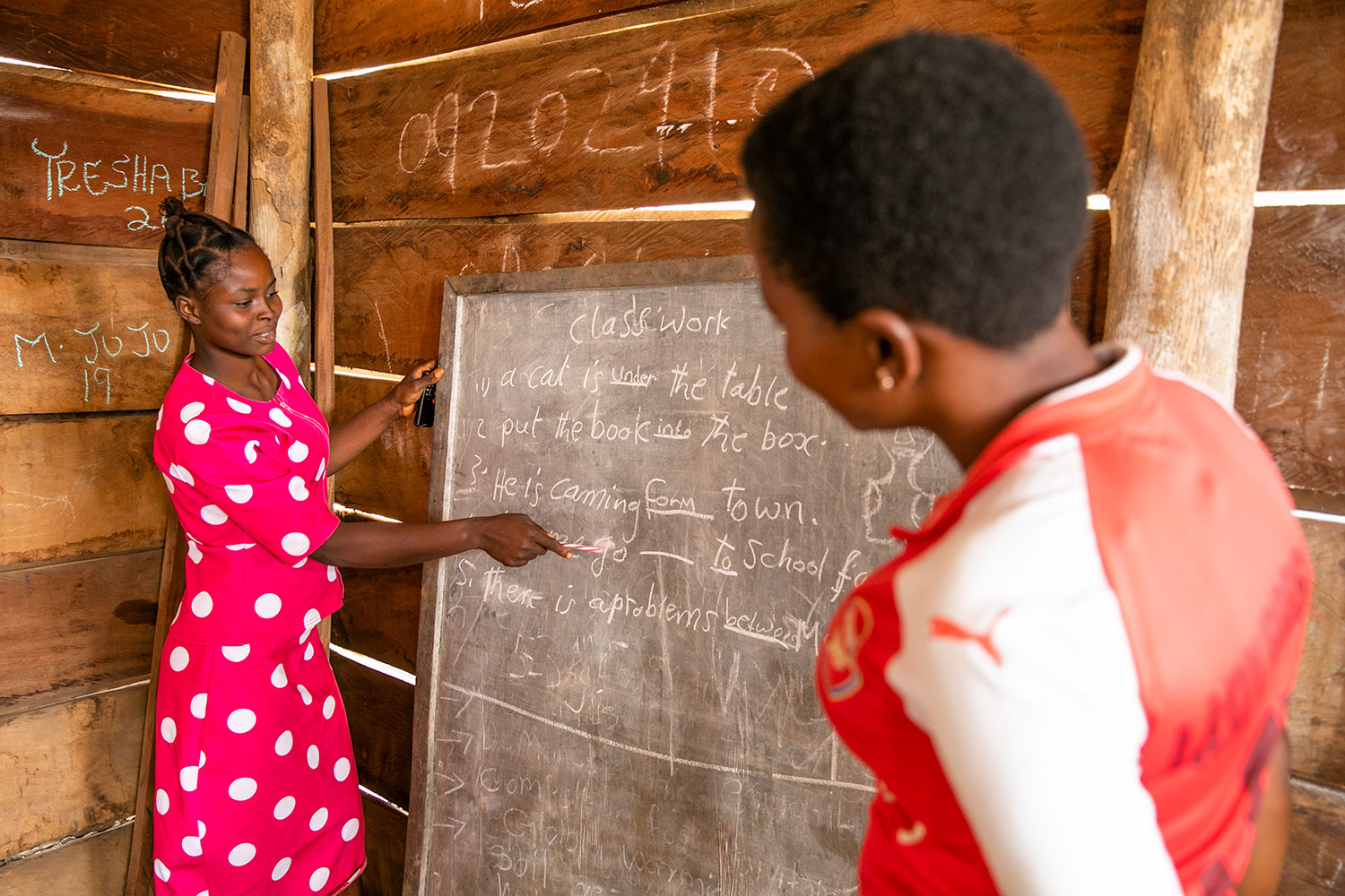 A teacher writes on a chalkboard as a young woman looks on