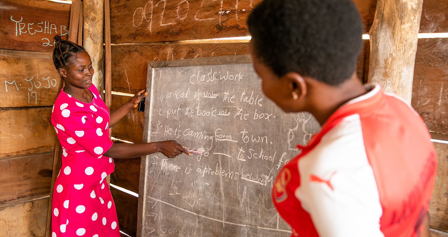 A teacher writes on a chalkboard as a young woman looks on