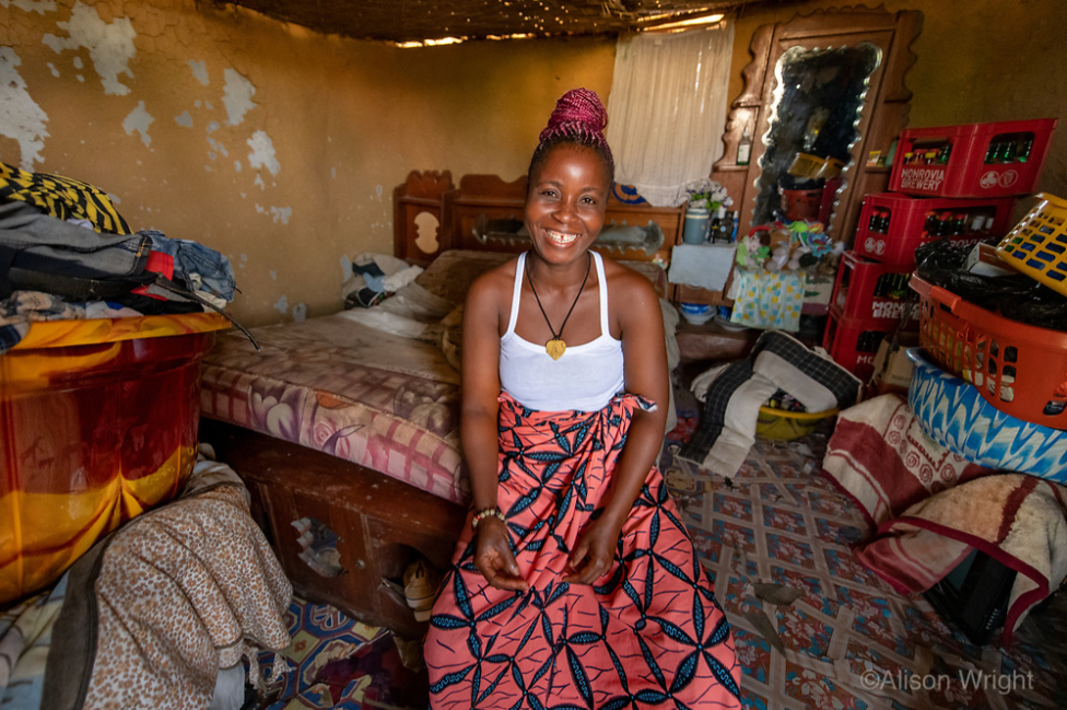 Jebbeh sits on the bed in her current home, a small two-room mud hut. Photo by Alison Wright.