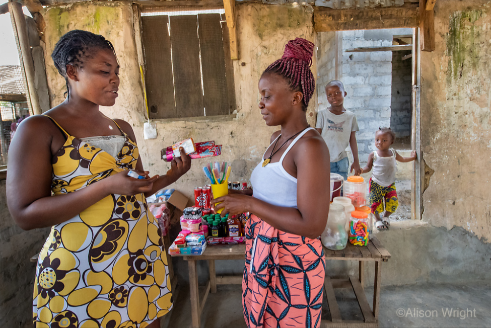 Jebbeh works with a customer, holding toothbrushes, in her new shop building while her two sons look on from behind. Photo by Alison Wright.