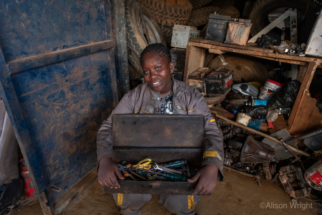 Josephine sits outside a tool shed holding a box of tools for fixing cars. Photo by Alison Wright.