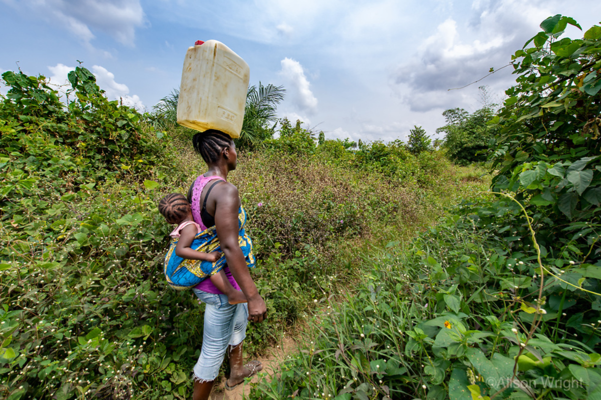 Patience carries a jug of water home from the fields with her baby on her back