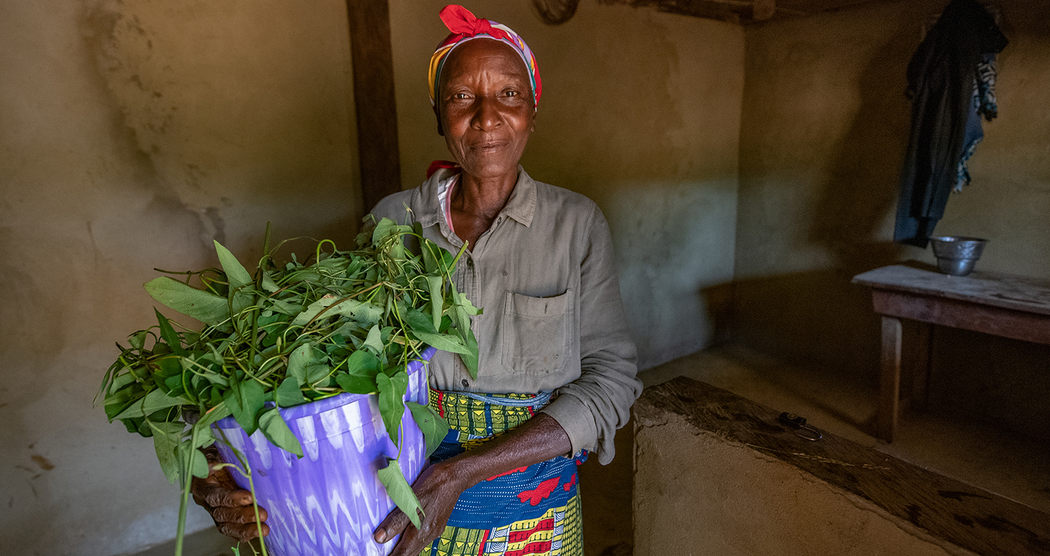 A woman in BRAC's agriculture program in Liberia poses in her home while holding a bucket of greens from her garden.