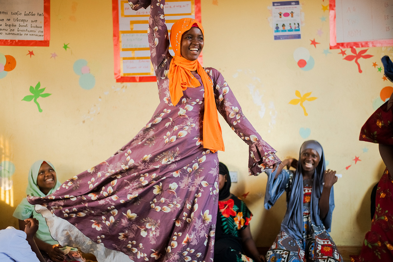 A smiling girl dances while her peers watch, clap, and laugh in a girls' empowerment club in Tanzania.