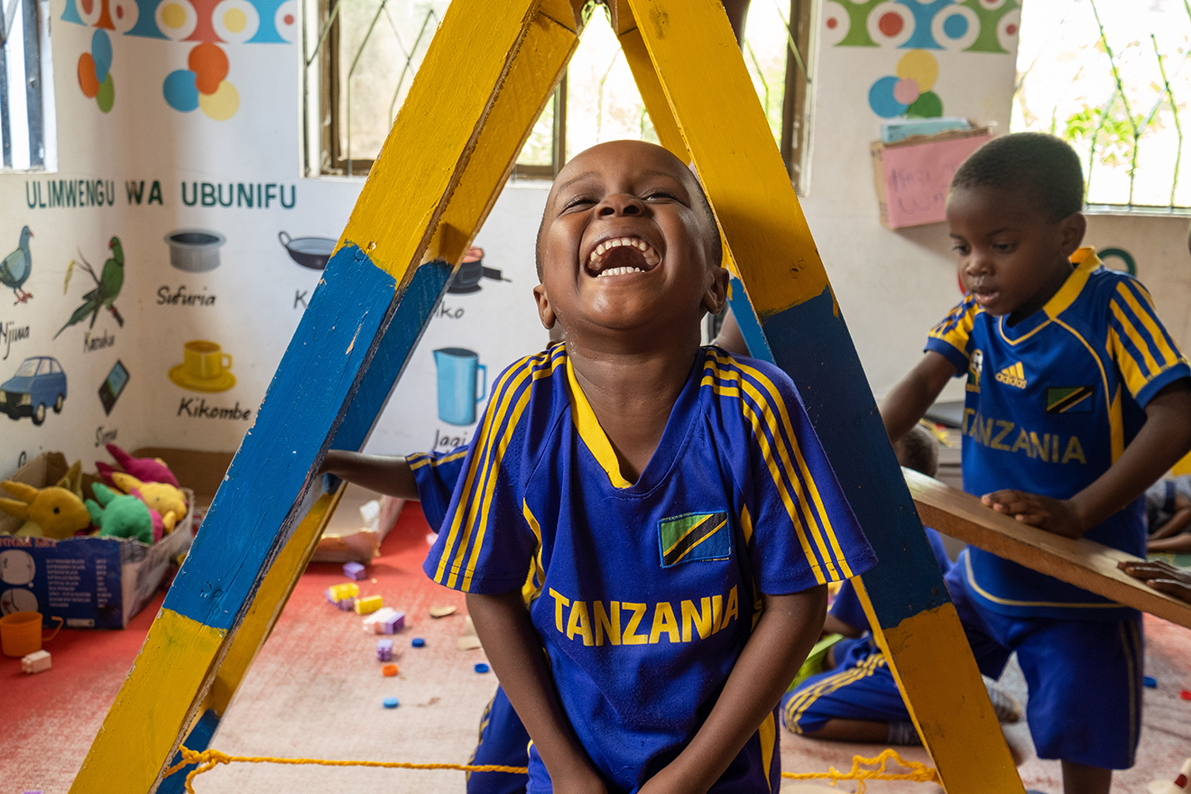 A child in a Play Lab wearing a Tanzania soccer jersey laughs while looking at the camera. Photo by Lee Cohen.