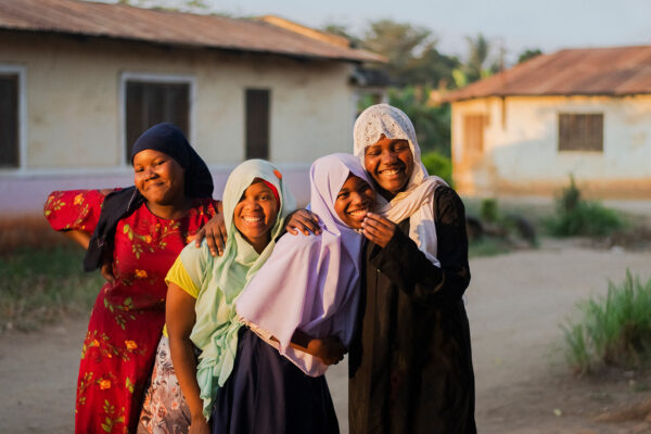 Four young women laugh while posing together outside of houses in a community in Tanzania.