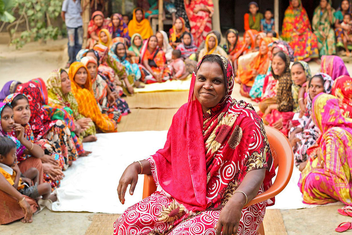 One woman leading a community meeting with BRAC smiles while surrounded by dozens of women from the community in Bangladesh