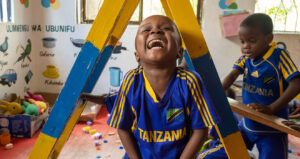 A child in a Play Lab laughs while looking at the camera and wearing a Tanzania soccer jersey. Photo by Lee Cohen.