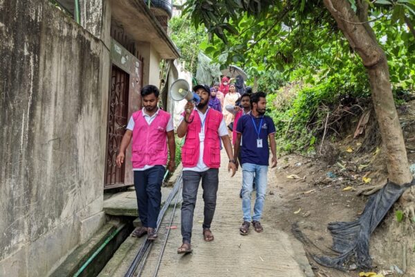 Three men walk down a dirt path alerting residents nearby to the dangers of the approaching storm. One man holds a megaphone.