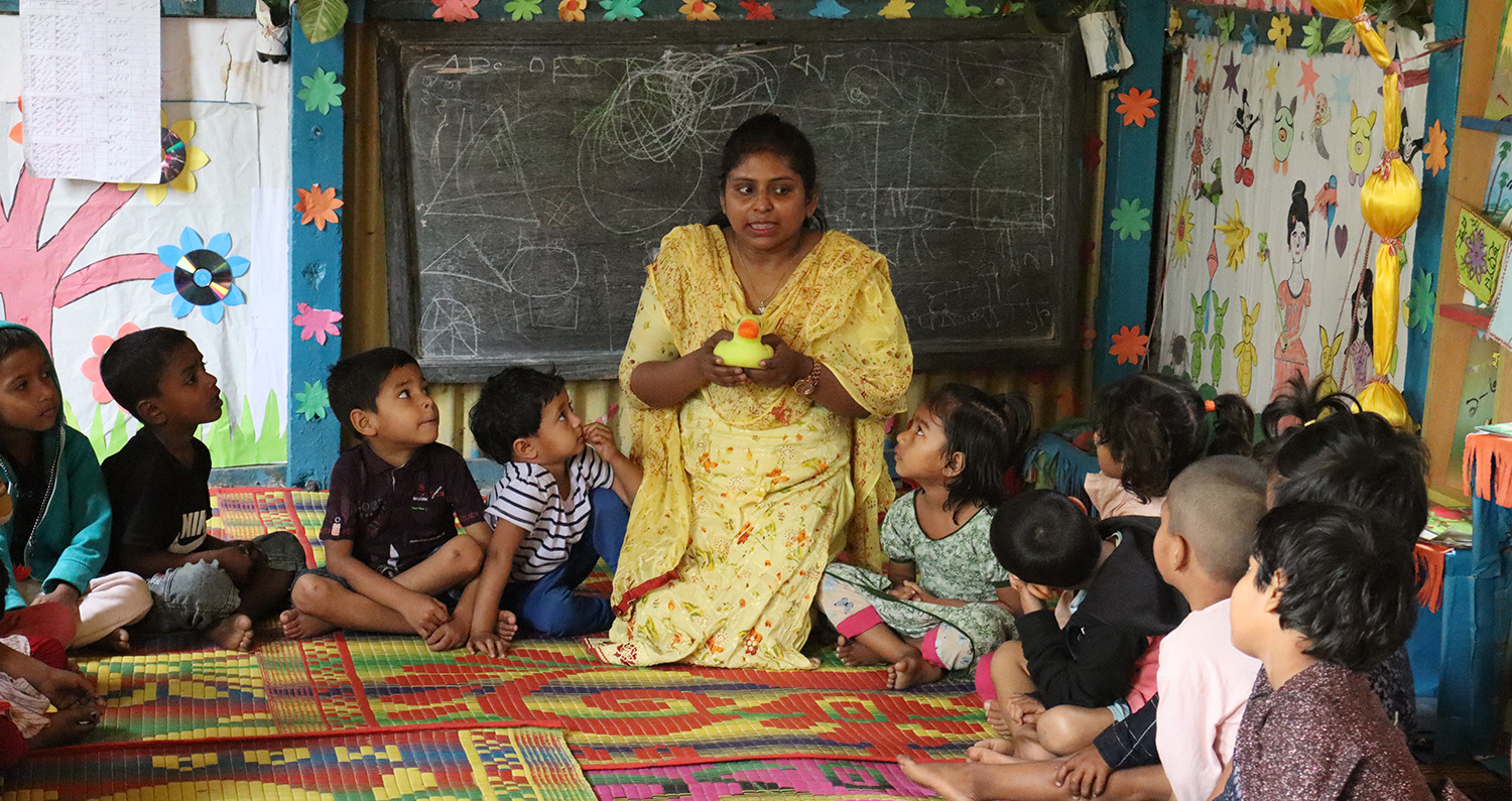 Amena, a Play Leader in a Play Lab in Dhaka, describes an activity holding a rubber duck while children sit in a circle and listen.
