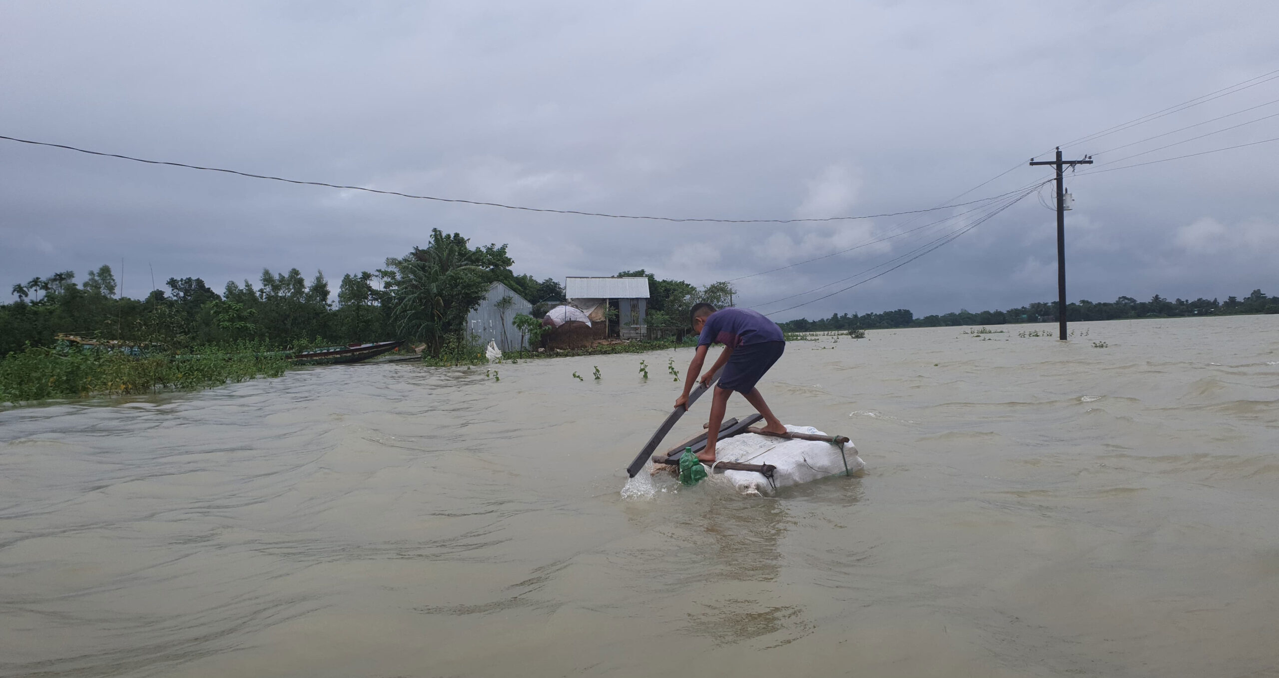 Under gray skies, a man leans forward on a makeshift raft to navigate flooding
