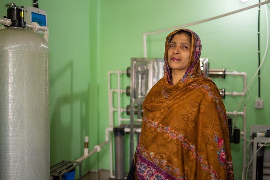 A clean water entrepreneur in Bangladesh poses next to her water filtration equipment and a large water tank.