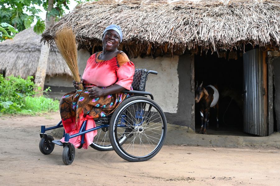 A woman who participated in a disability-inclusive Graduation program in Uganda poses in front of a small hut where her goats live, while sitting in her wheelchair.