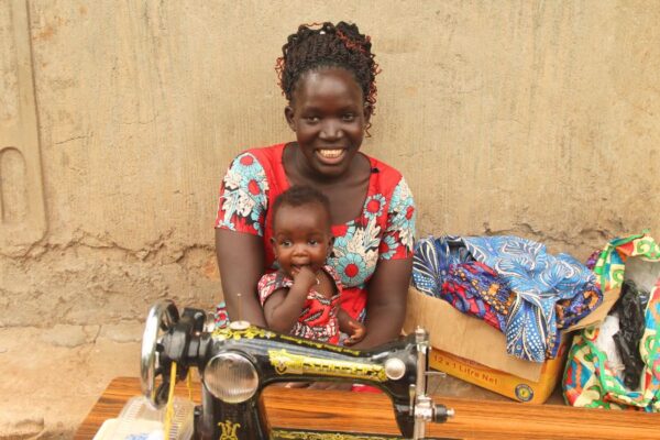 Lonah poses with her sewing machine while her small baby sits in her lap. They both wear vibrant red dresses that she sewed.