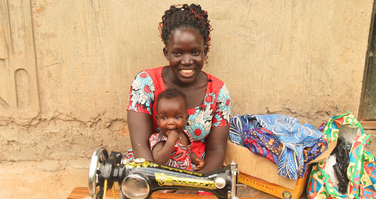 Lonah poses with her sewing machine while her small baby sits in her lap. They both wear vibrant red dresses that she sewed.