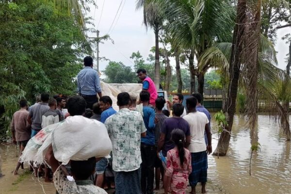 A crowd of about a dozen people stand in calf-deep behind a truck. They wait to collect emergency food items in Sylhet. One man in the foreground has a white bag on his head.