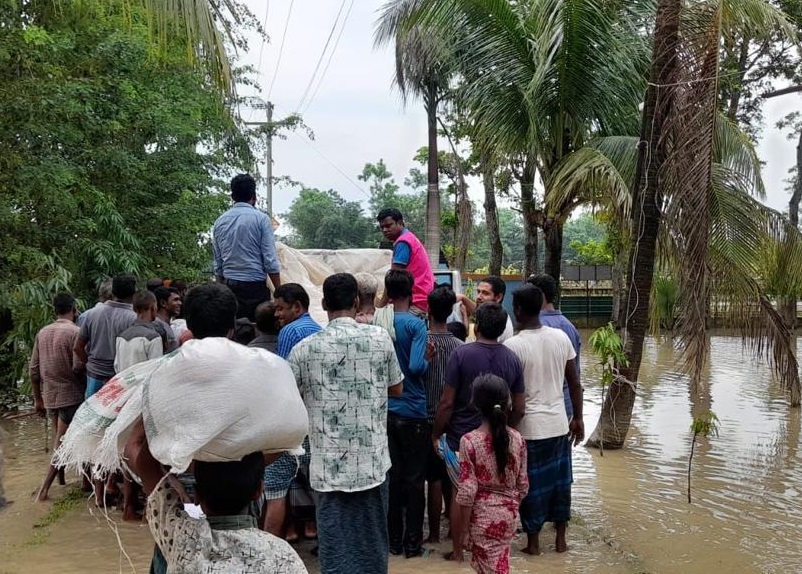 A crowd of about a dozen people stand in calf-deep behind a truck. They wait to collect emergency food items in Sylhet. One man in the foreground has a white bag on his head.