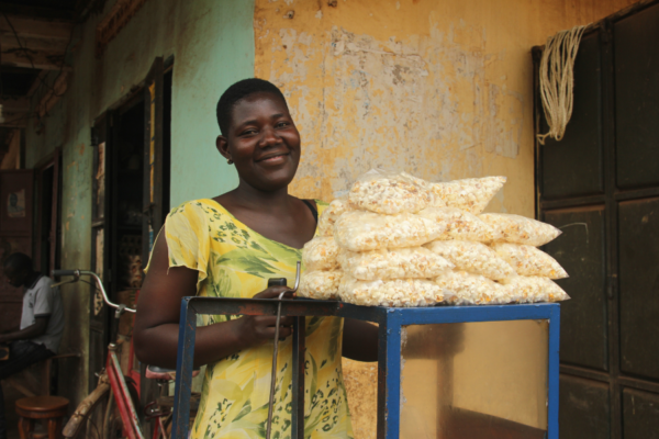 Juliet, a young woman in BRAC's youth empowerment program in Uganda, smiles as she poses behind her popcorn machine with a pile of bags of popcorn perched atop for sale.