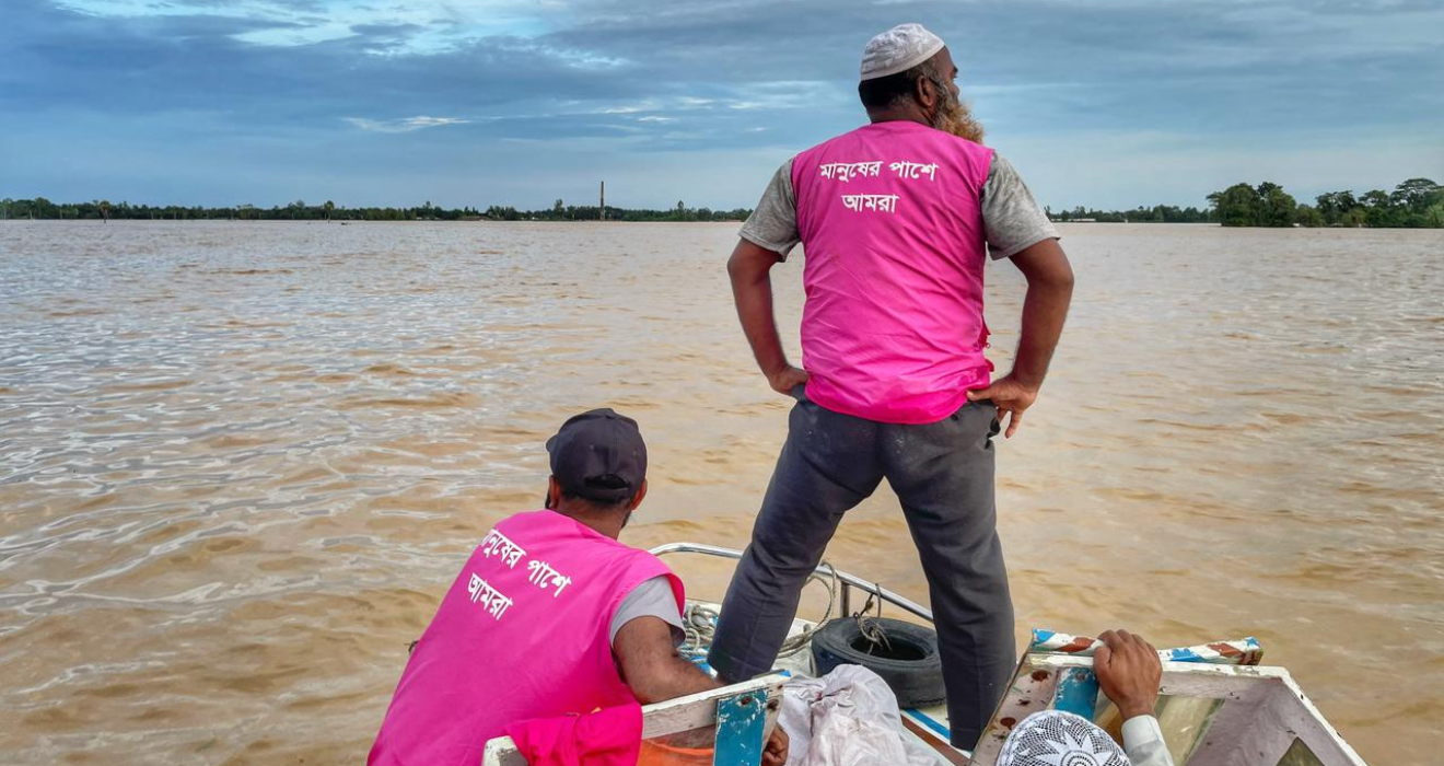 Two BRAC staffers wearing magenta BRAC vests look out ahead over the floodwaters while traveling via boat to deliver aid. 