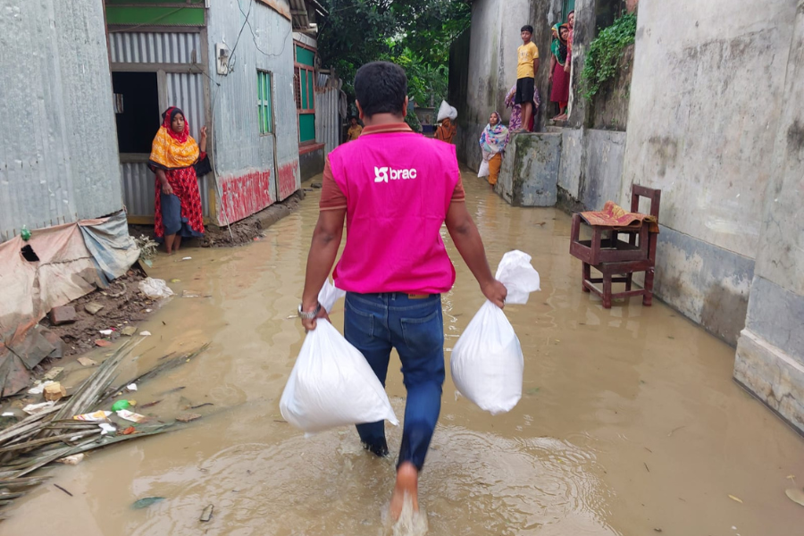 A BRAC staff member carrying two large sacks of food aid wades through a flooded alley.