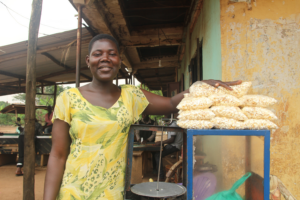 Juliet, a young woman in BRAC's youth empowerment program in Uganda, smiles as she poses behind her popcorn machine with a pile of bags of popcorn perched atop for sale.