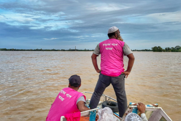 Two BRAC staffers wearing magenta BRAC vests look out ahead over the floodwaters while traveling via boat to deliver aid.