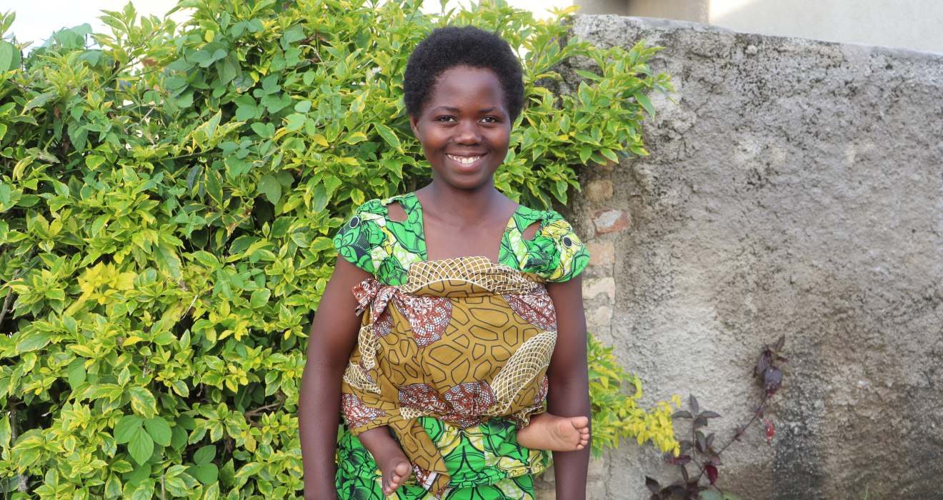 Angelique poses outside in front of greenery with her baby on her back.