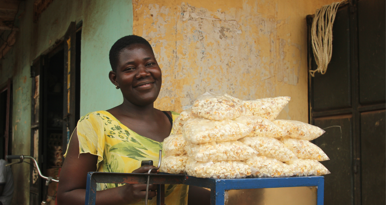 Juliet, a young woman in BRAC's youth empowerment program in Uganda, smiles as she poses behind her popcorn machine with a pile of bags of popcorn perched atop for sale.