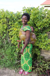 Angelique poses outside in front of greenery with her baby on her back.