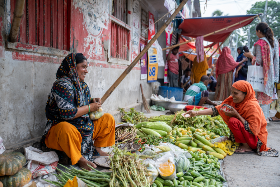 A woman in BRAC's Ultra-Poor Graduation program sits at her market produce stall and smiles at a shopping customer.