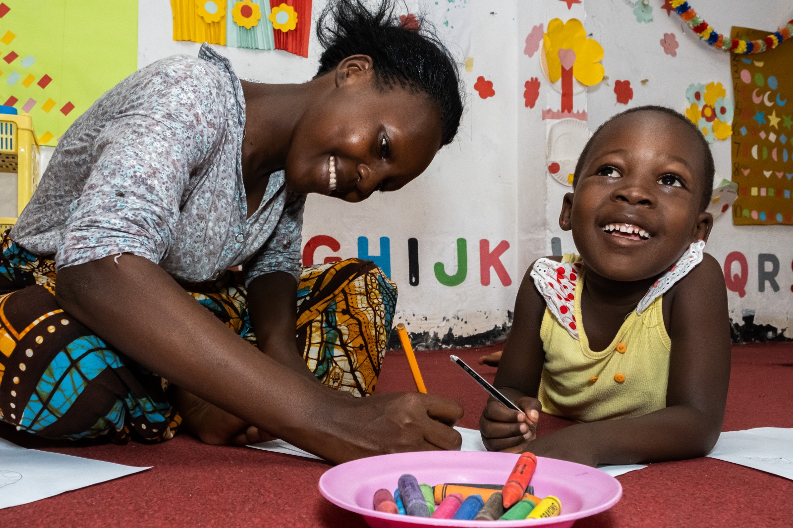 A smiling woman in a white shirt sits on the floor, coloring alongside a young child in a yellow top. The child is looking up and is smiling. There is a pink plate in front of them with orange, green and yellow crayons.