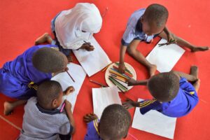 Six children sit on a pink floor, each with white pieces of paper and a stack of colored pencils with which to draw. 