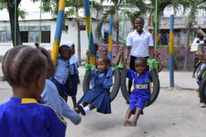 Two children wearing blue school uniforms sit on tire swings. A smiling teacher wearing a white shirt stands behind them. 