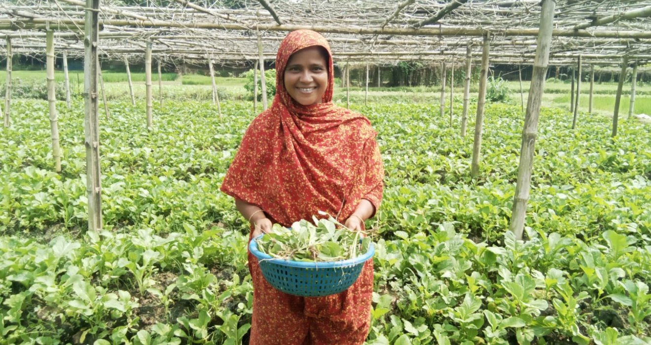 An image of a woman, Rozina, standing in her farm with crops behind her and holding harvested leafy greens.