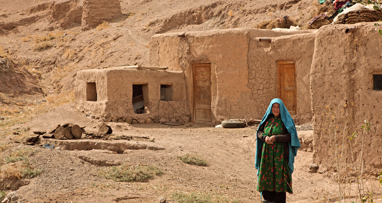 A woman stands in front of a home made of mud bricks in Afghanistan.