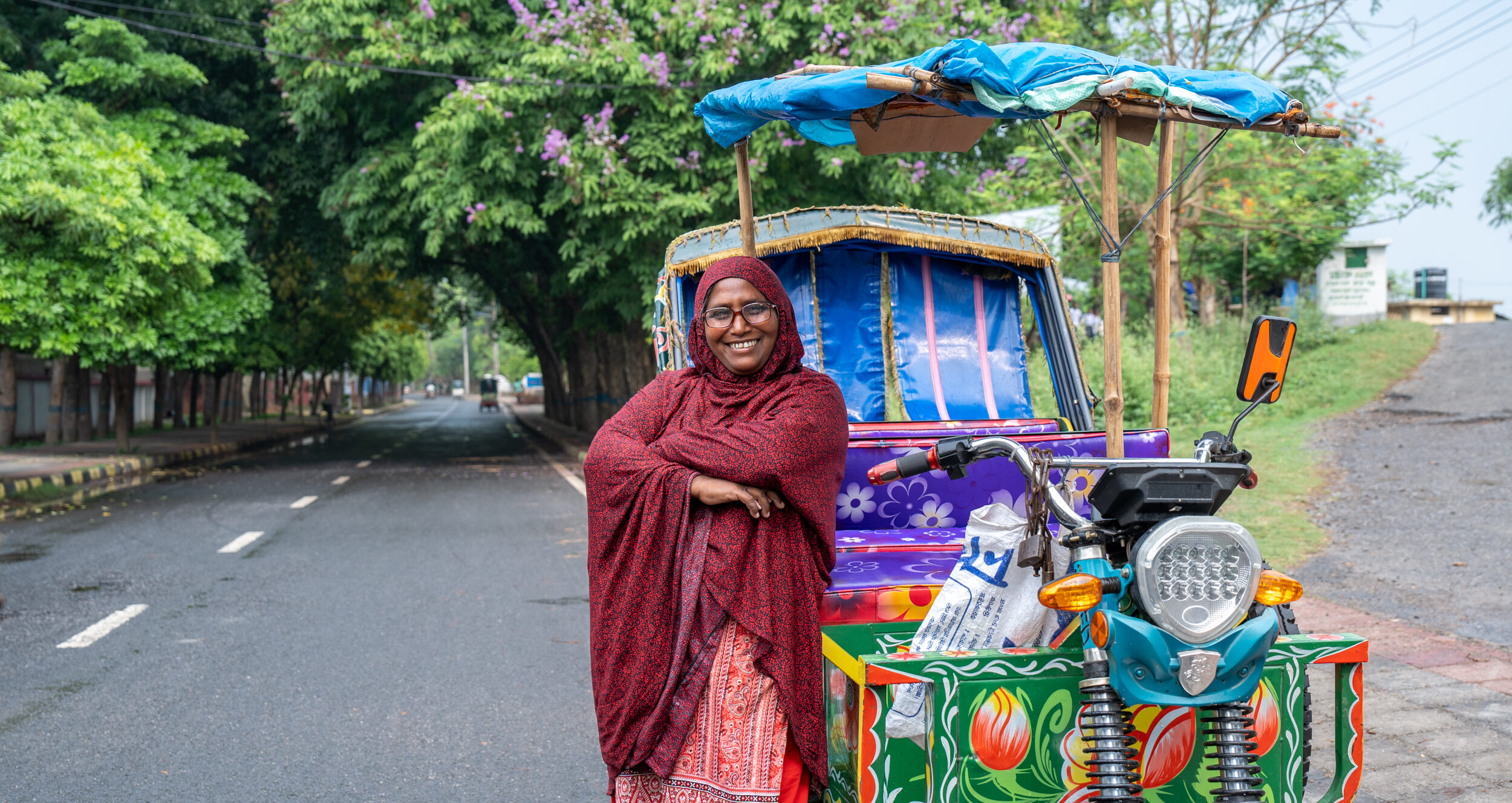 A woman poses with her arms crossed proudly while leaning against her tuk tuk on a road in Bangladesh.