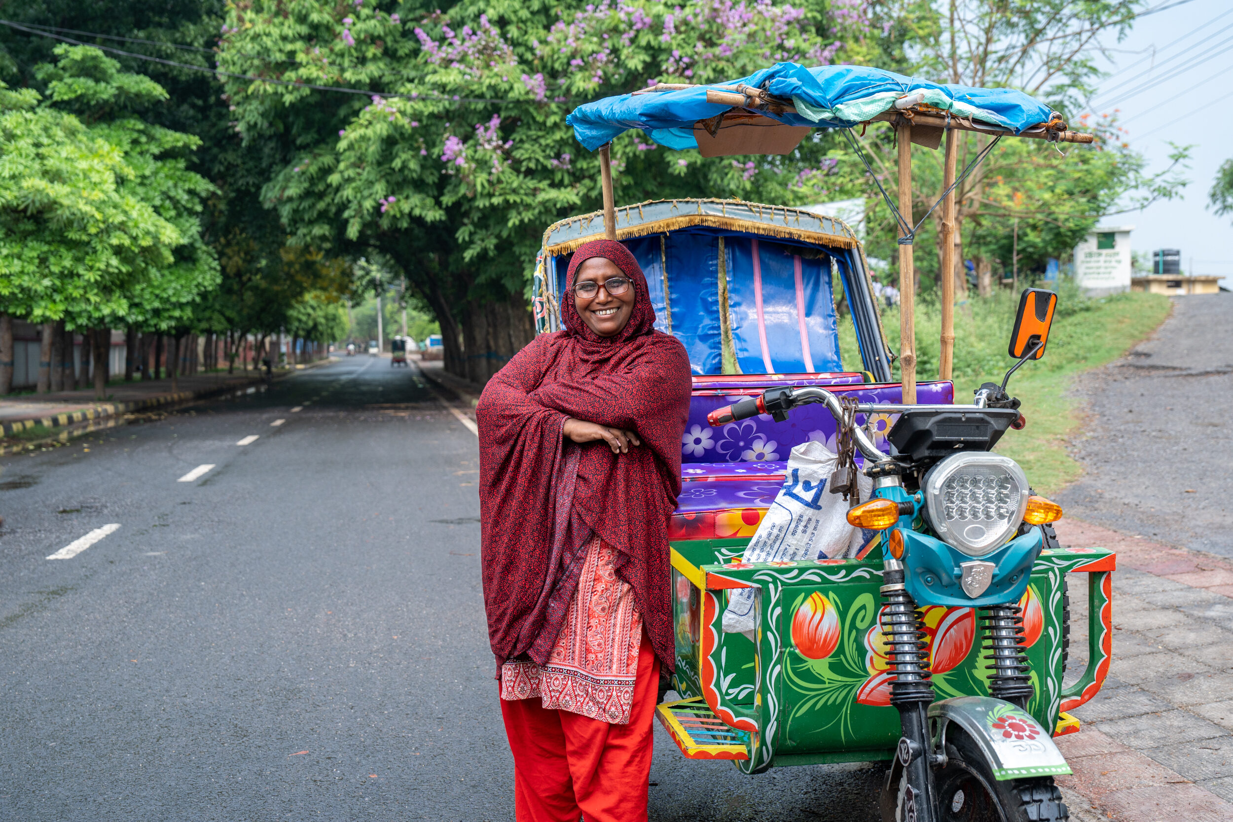 A woman poses with her arms crossed proudly while leaning against her tuk tuk on a road in Bangladesh.