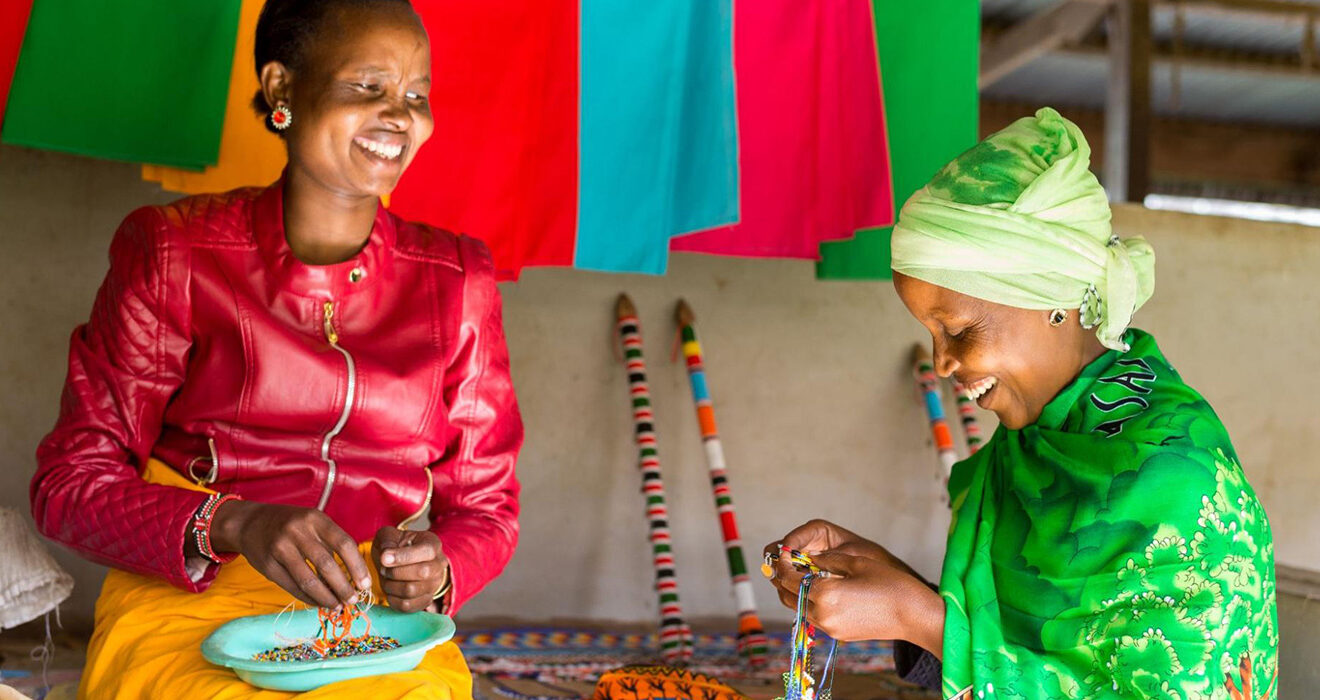 Two women in Kenya smile together as they make beaded jewelry in front of a backdrop of colorful fabrics hanging on the wall of a simple shop.