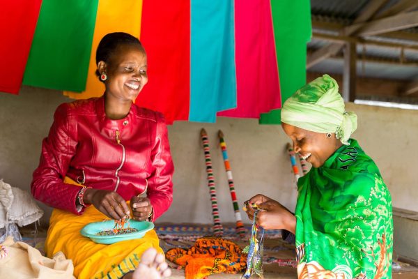 Two women in Kenya smile together as they make beaded jewelry in front of a backdrop of colorful fabrics hanging on the wall of a simple shop.