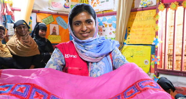 A woman in a BRAC vest stands in a BRAC safe space while holding up a pink piece of fabric with embroidery on it.
