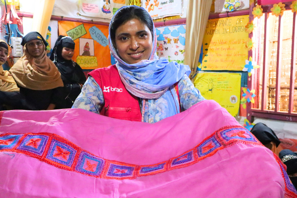 A woman in a BRAC vest stands in a BRAC safe space while holding up a pink piece of fabric with embroidery on it.