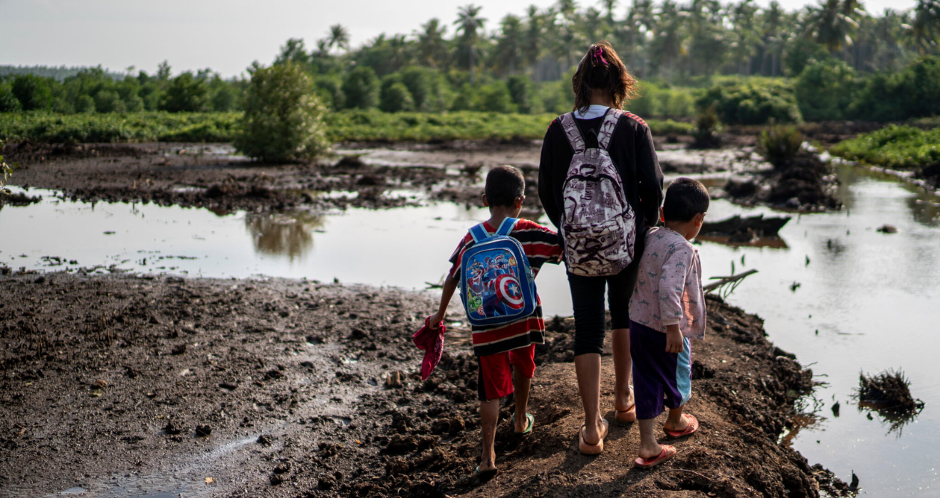 Two girls and a boy hold hands while walking to school wearing backpacks, as they overlook a waterway and forest that they must cross.