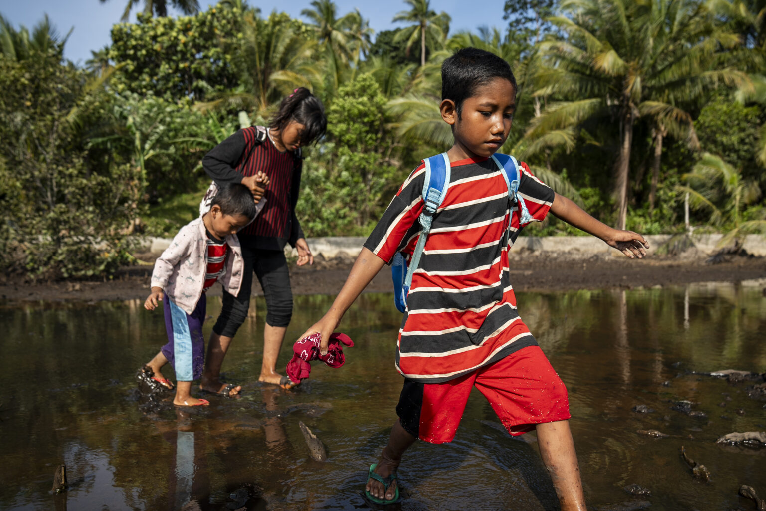 Two girls and one boy trapse through standing water while wearing backpacks on the way to school in the Philippines.
